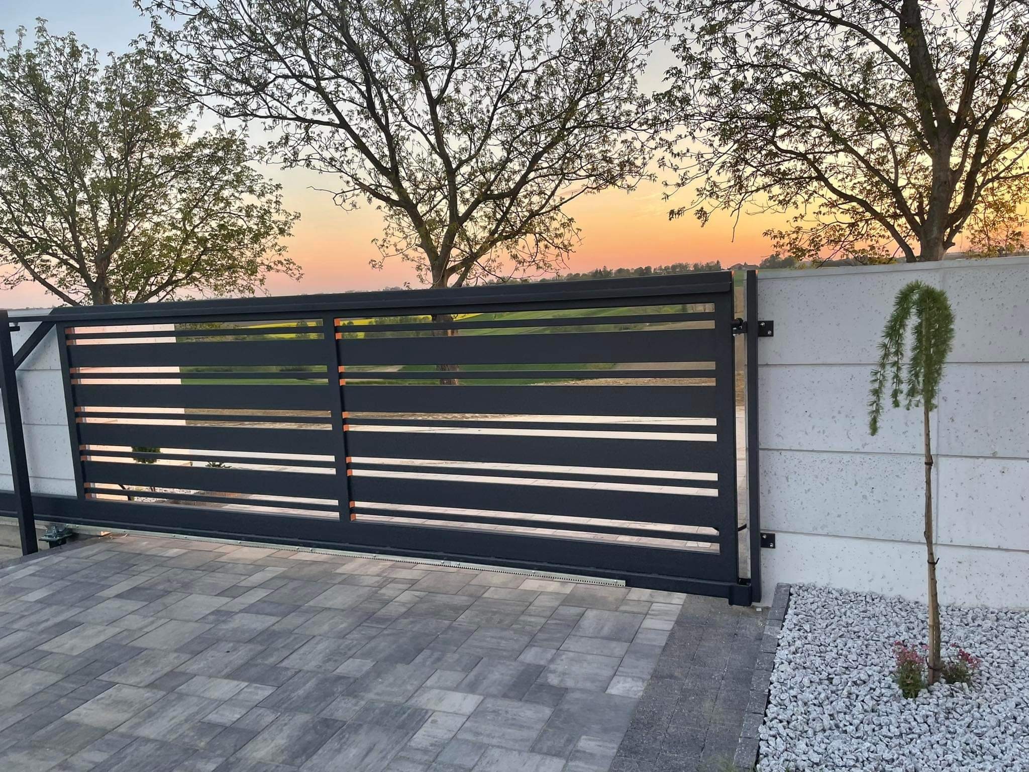 Nowoczesna, przesuwna brama wjazdowa z poziomymi panelami na tle zachodzącego słońca i dekoracyjnej roślinności.