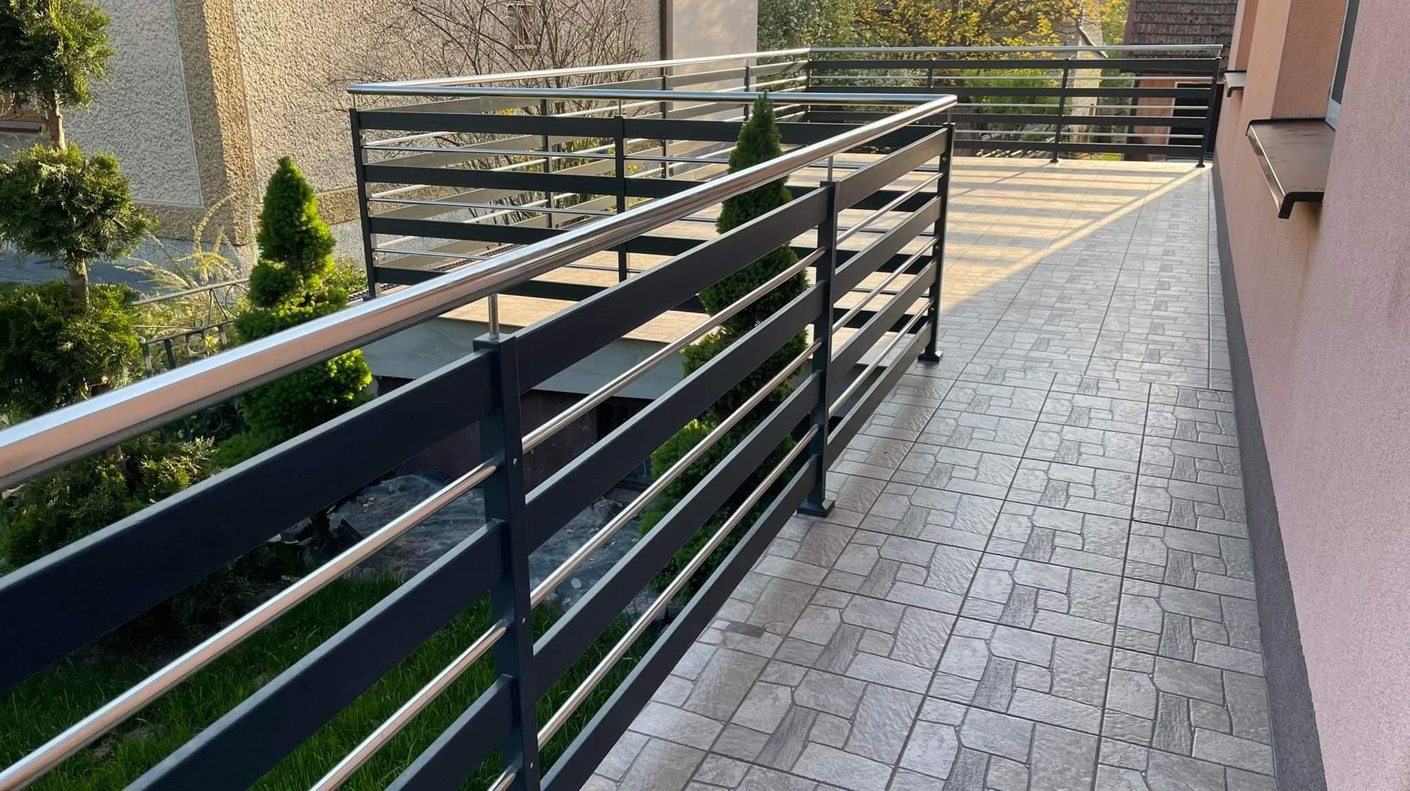 Nowoczesna balustrada balkonowa z elementami stali nierdzewnej i ciemnoszarego metalu, widok z boku, z widocznymi elementami roślinności w tle.