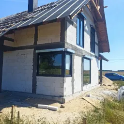 Budowa domu jednorodzinnego z zamontowanymi oknami PCV i roletami zewnętrznymi, widoczne prace wykończeniowe elewacji i dachu, w tle błękitny samoch&oacute;d.