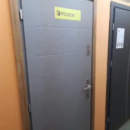 Szare drzwi z drewnianą ramą i naklejoną żółtą etykietą producenta Polstar, prezentowane w sklepie obok ciemniejszych drzwi.