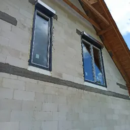 Montaż okien z roletami zewnętrznymi w surowym stanie budynku z betonu kom&oacute;rkowego, widoczne pianowanie i częściowe wykończenie elewacji.