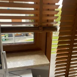 Drewniane żaluzje poziome w kolorze miodowym, częściowo odsłonięte, z widokiem na podwórze z zielonym trawnikiem i koszami na śmieci przez okno o drewnianej ramie.