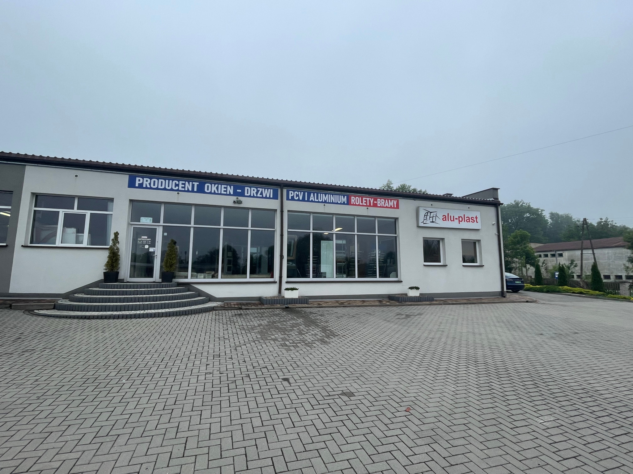Fasada budynku firmy Alu-plast z dużymi oknami wystawowymi, prezentująca ofertę okien, drzwi, rolet i bram. Na budynku widoczne logo firmy.