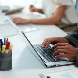 Pracownik biurowy w szarej marynarce pisze na laptopie, obok kubek z pisakami i dokumenty z wykresami, w tle druga osoba przy biurku.