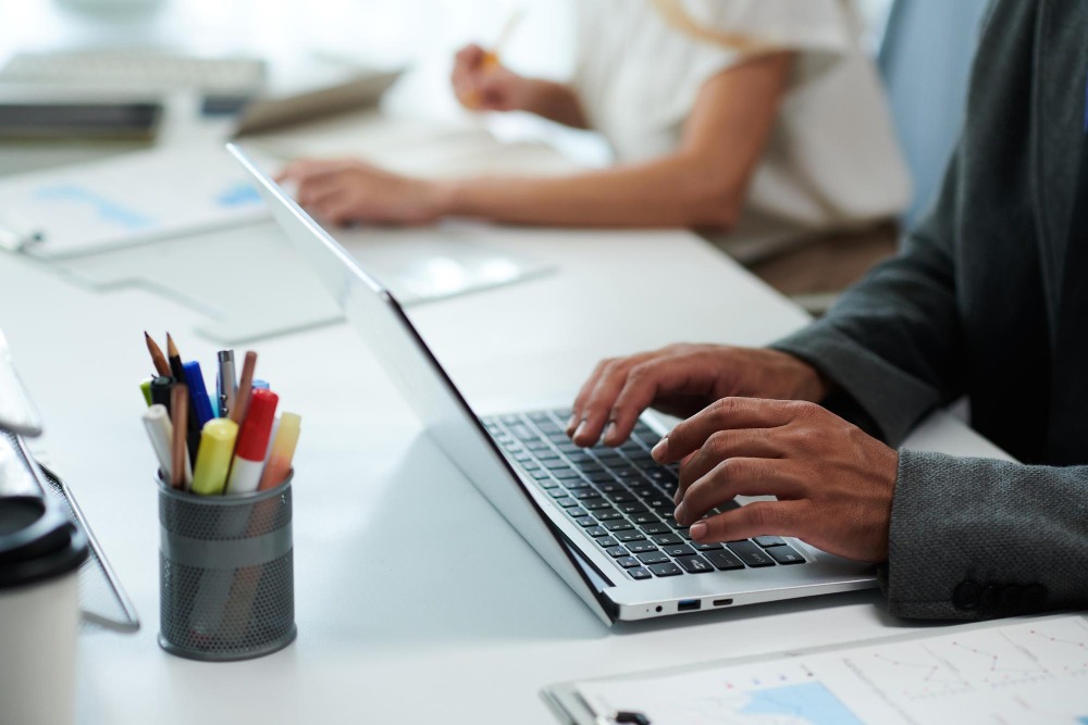 Pracownik biurowy w szarej marynarce pisze na laptopie, obok kubek z pisakami i dokumenty z wykresami, w tle druga osoba przy biurku.