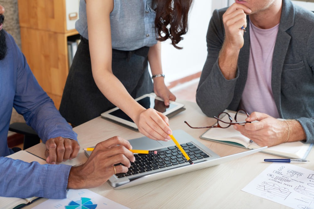 Zespół analizuje dane na laptopie, używając ołówków do wskazywania kluczowych informacji na ekranie, w tle tablet i notatnik.
