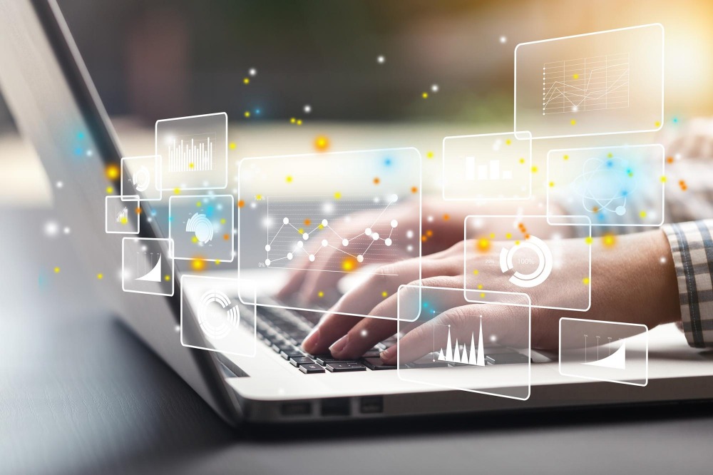 Osoba wpisuje dane na laptopie z nałożonymi grafikami wykresów i wskaźników, sugerując analizę finansową lub raportowanie.