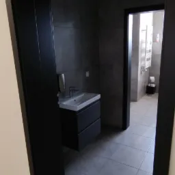 Nowoczesna łazienka z szarą armaturą i czarnymi futrynami drzwi, widoczna umywalka z szafką, szare płytki podłogowe i fragment grzejnika w dalszej części pomieszczenia.