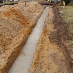 Wykop fundamentowy wypełniony świeżym betonem, widoczne pryzmy piasku i ziemi, w tle elementy drewnianej konstrukcji.