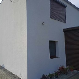 APM Marcin Woś Ambasador Rockwoll - Fragment domu z jasną elewacją, brązowymi roletami zewnętrznymi i małym oknem, oświetlony lampą zewnętrzną, z dekoracyjnym kamieniem i roślinami wzdłuż ściany.