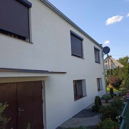 APM Marcin Woś Ambasador Rockwoll - Biały dom jednorodzinny z brązowymi roletami zewnętrznymi i garażem, widok z boku na elewację i zadbany ogródek przydomowy z kamykami.