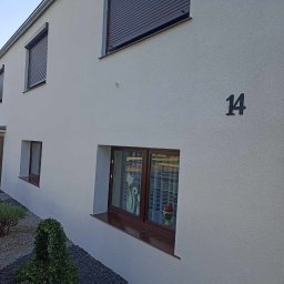 APM Marcin Woś Ambasador Rockwoll - Elewacja domu jednorodzinnego o jasnej, chropowatej strukturze, z ciemnymi roletami i numerem '14' na ścianie, widoczny fragment ogrodu z kamieniami i roślinami.