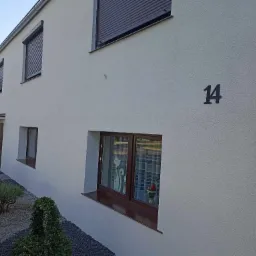 Elewacja domu jednorodzinnego o jasnej, chropowatej strukturze, z ciemnymi roletami i numerem '14' na ścianie, widoczny fragment ogrodu z kamieniami i roślinami.