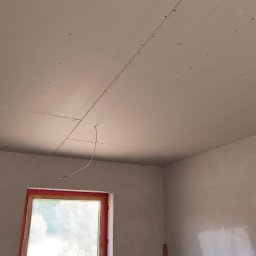 APM Marcin Woś Ambasador Rockwoll - Montaż płyt karton-gips na suficie w pokoju z oknem. Widoczny podnośnik do płyt i fragmenty ścian z lekkimi niedoskonałościami. Etap prac wykończeniowych.