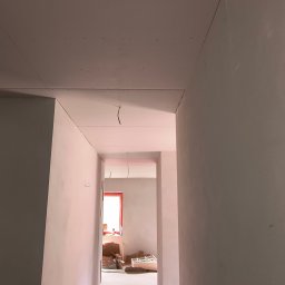 APM Marcin Woś Ambasador Rockwoll - Korytarz w trakcie prac wykończeniowych: zamontowane płyty karton-gips na ścianach i suficie, widoczne kable. Czerwona rama okna w oddali.