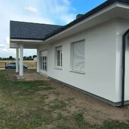 Parterowy dom jednorodzinny z białymi ścianami i ciemnym dachem. Widoczne rolety zewnętrzne, trawnik i fragment nieba. Ujęcie z boku, podkreślające bryłę budynku.