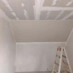 Poddasze w trakcie wykańczania z widocznymi płytami kartonowo-gipsowymi na suficie, częściowo wyszpachlowanymi, z wystającym przewodem elektrycznym i drewnianą drabiną w rogu pomieszczenia.