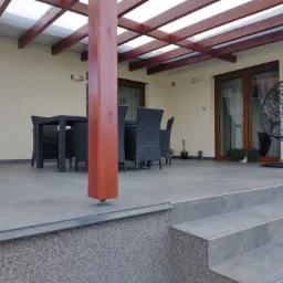 Zadaszony taras z szarych płytek, drewniane słupy podtrzymujące zadaszenie, czarne meble ogrodowe i wiszący fotel, widok z dołu na schody tarasowe.