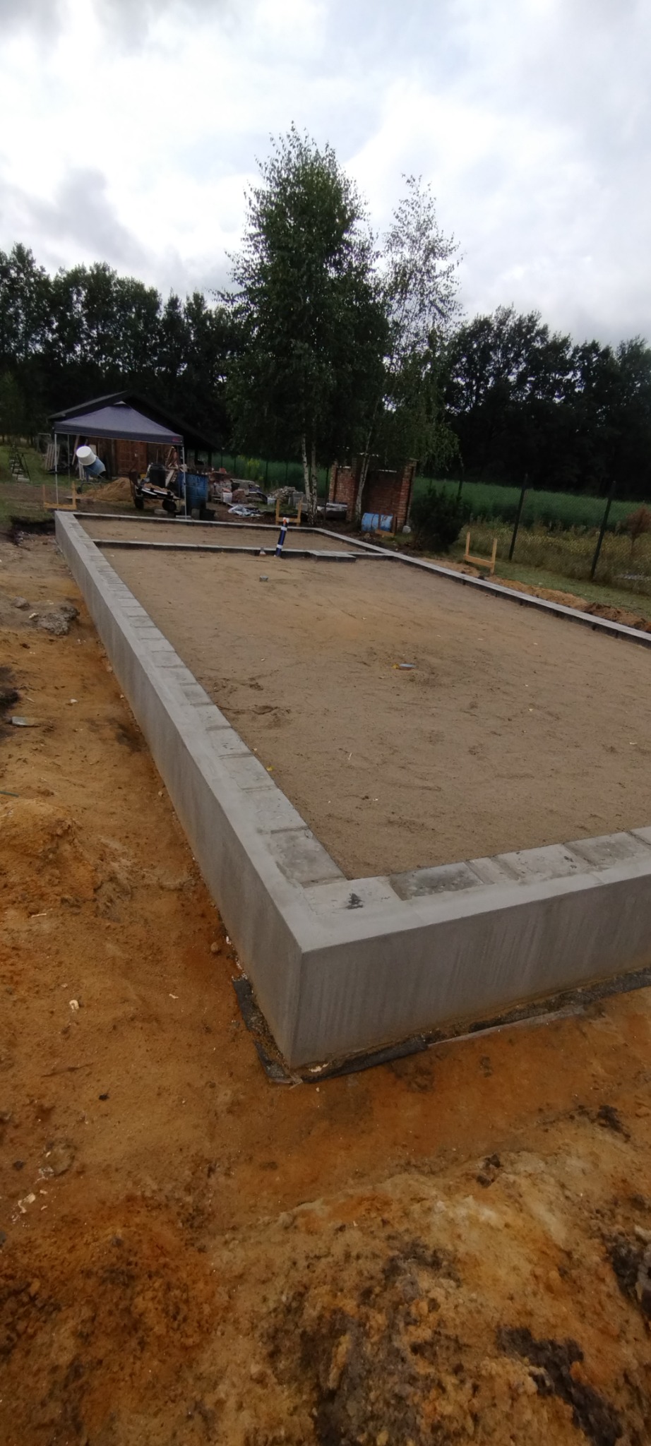 Wylany fundament pod budowę domu, widoczne szalunki i przygotowany teren, w tle drzewa i pochmurne niebo.