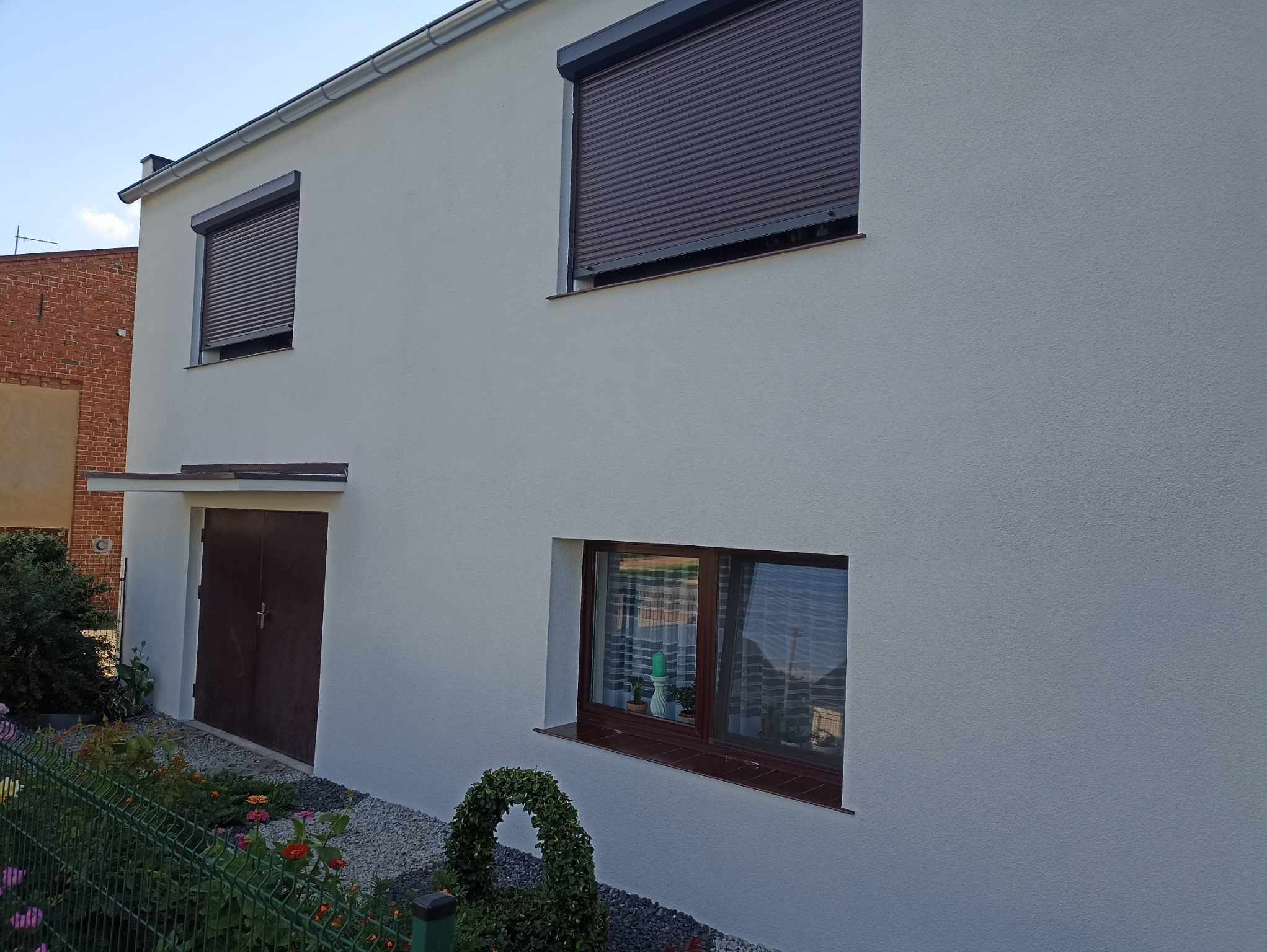 Jasnoszary dom jednorodzinny z brązowymi roletami i drzwiami garażowymi, świeżo po wykonaniu elewacji z widoczną strukturą tynku oraz zadbanym ogrodem z przodu.