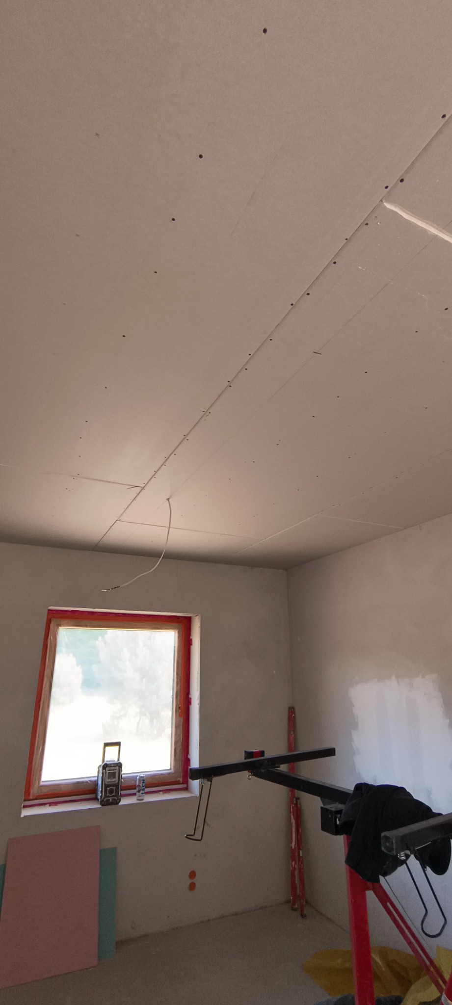 Montaż płyt karton-gips na suficie w pokoju z oknem. Widoczny podnośnik do płyt i fragmenty ścian z lekkimi niedoskonałościami. Etap prac wykończeniowych.