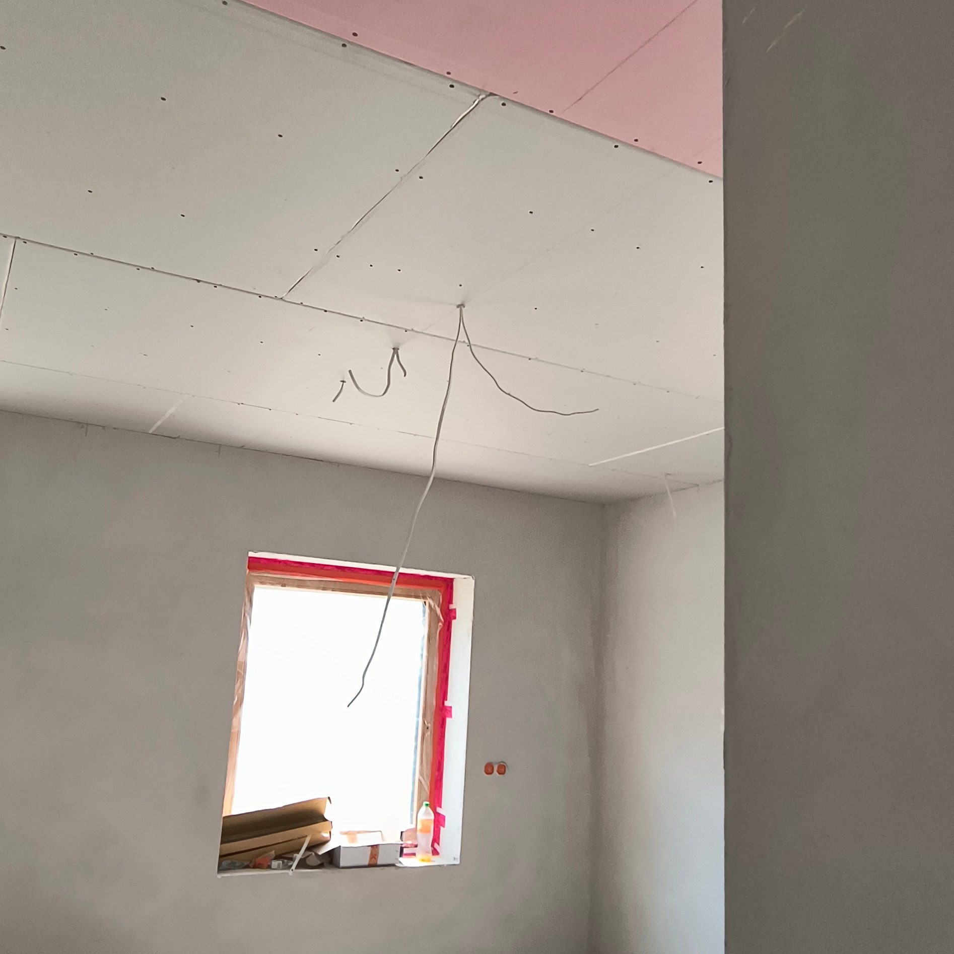 Wnętrze w trakcie prac: surowe ściany, okno z zabezpieczeniem, częściowo zamontowana płyta gipsowa na suficie z wystającymi przewodami.