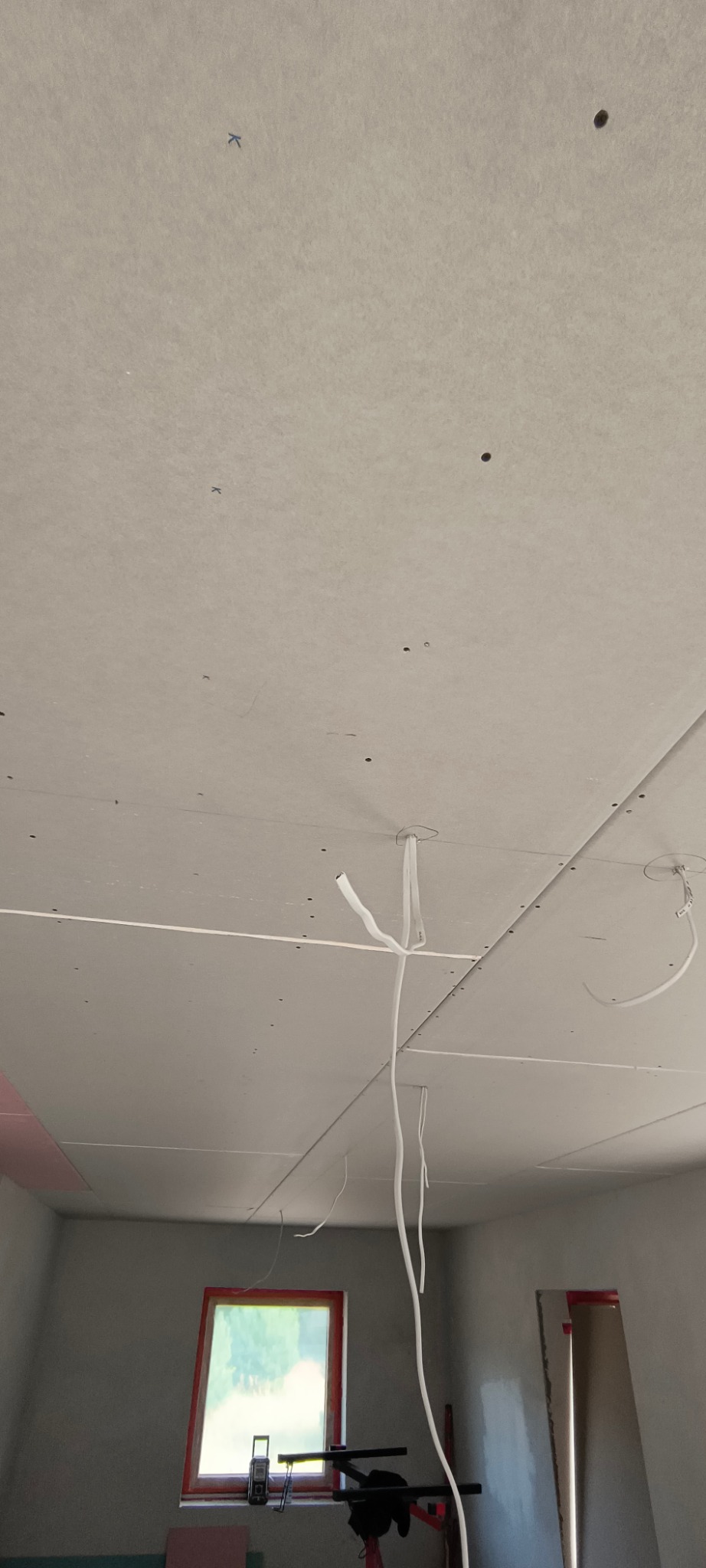 Sufit z płyt gipsowo-kartonowych w trakcie montażu, widoczne przewody elektryczne. Ujęcie z dołu, okno w tle. Ślady po szpachlowaniu.