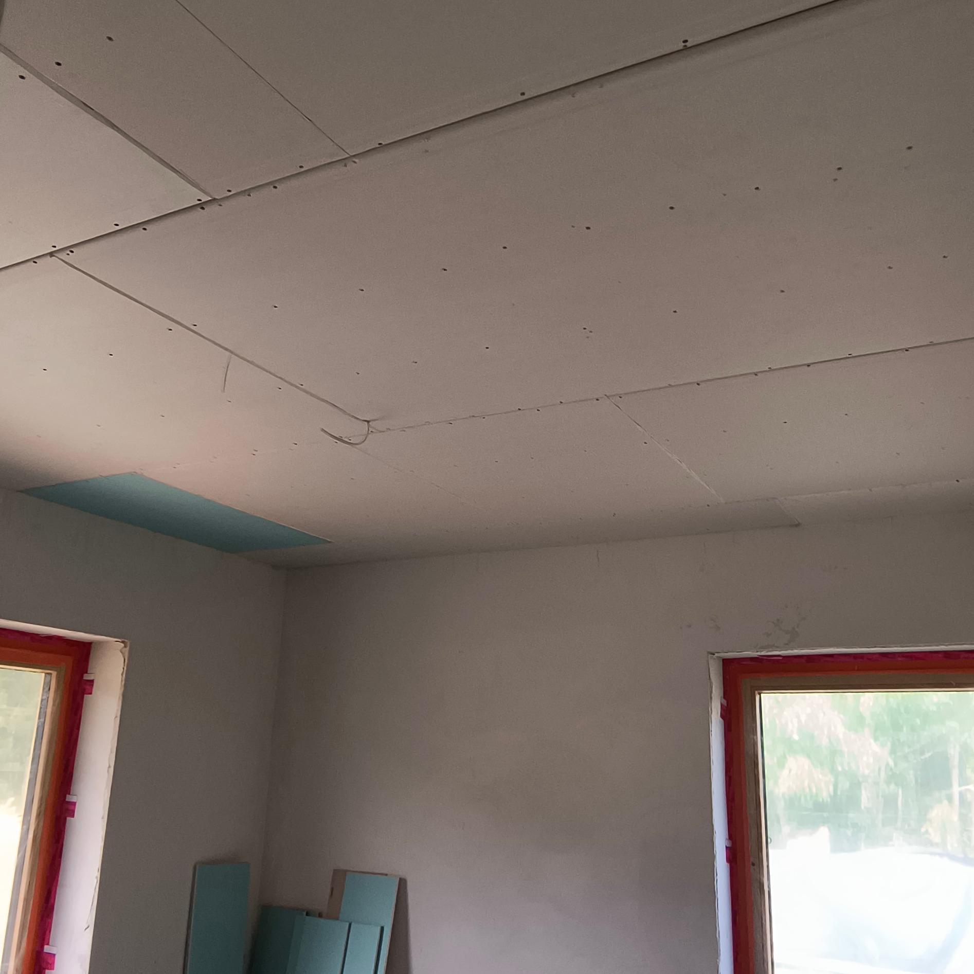 Montaż płyt gipsowo-kartonowych na suficie, widoczne łączenia i otwór rewizyjny. W tle okna z czerwoną folią ochronną i płyty g-k przy ścianie.