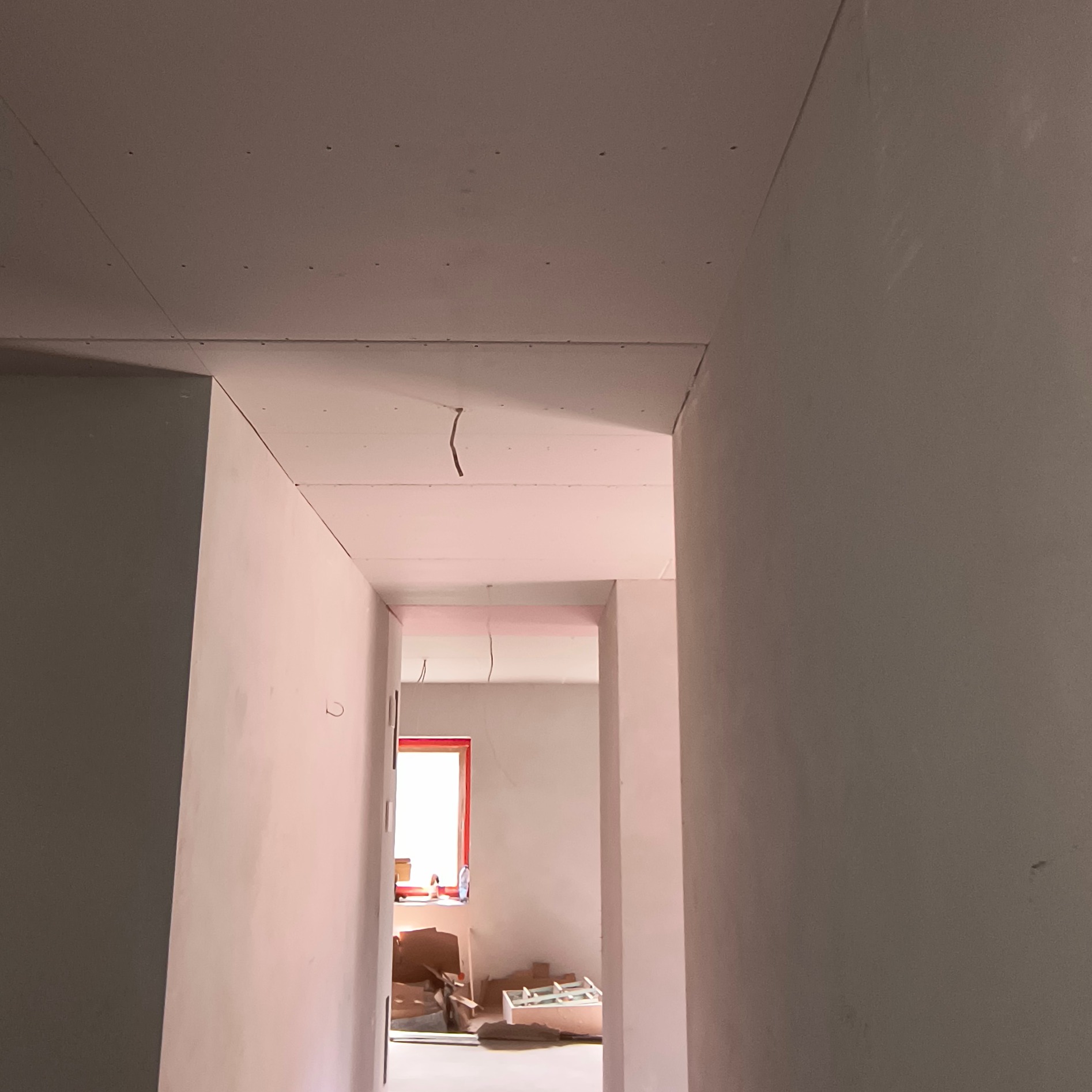 Korytarz w trakcie prac wykończeniowych: zamontowane płyty karton-gips na ścianach i suficie, widoczne kable. Czerwona rama okna w oddali.