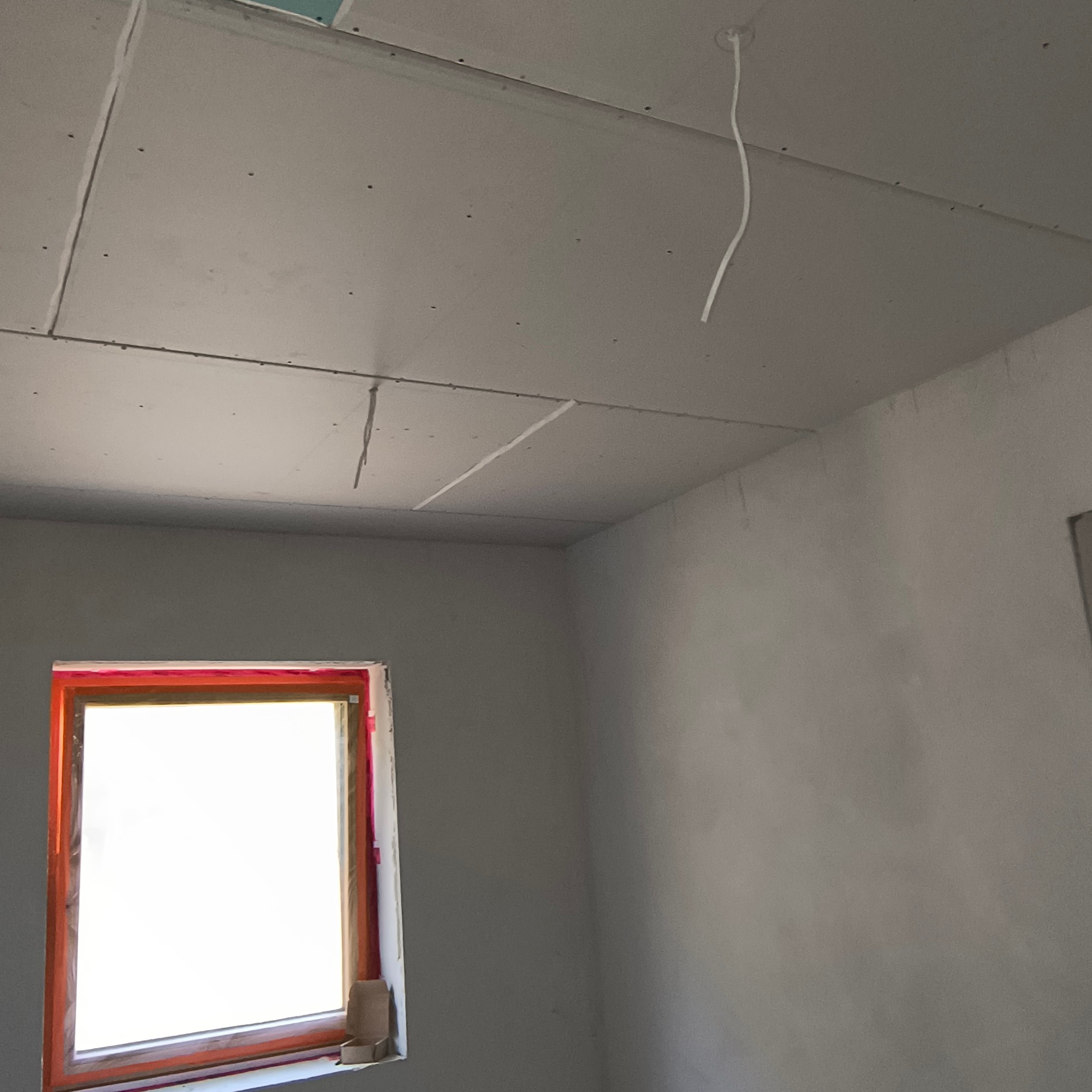 Montaż płyt karton-gips na suficie w Nowym Tomyślu. Widoczne łączenia płyt i wystające przewody. Okno zabezpieczone folią malarską.