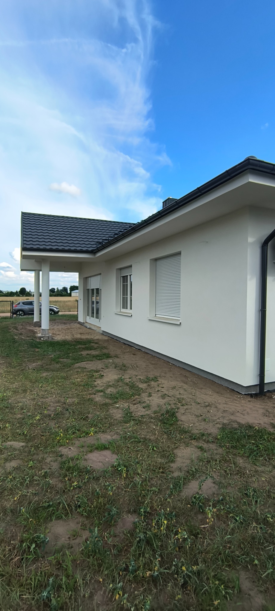 Parterowy dom jednorodzinny z białymi ścianami i ciemnym dachem. Widoczne rolety zewnętrzne, trawnik i fragment nieba. Ujęcie z boku, podkreślające bryłę budynku.