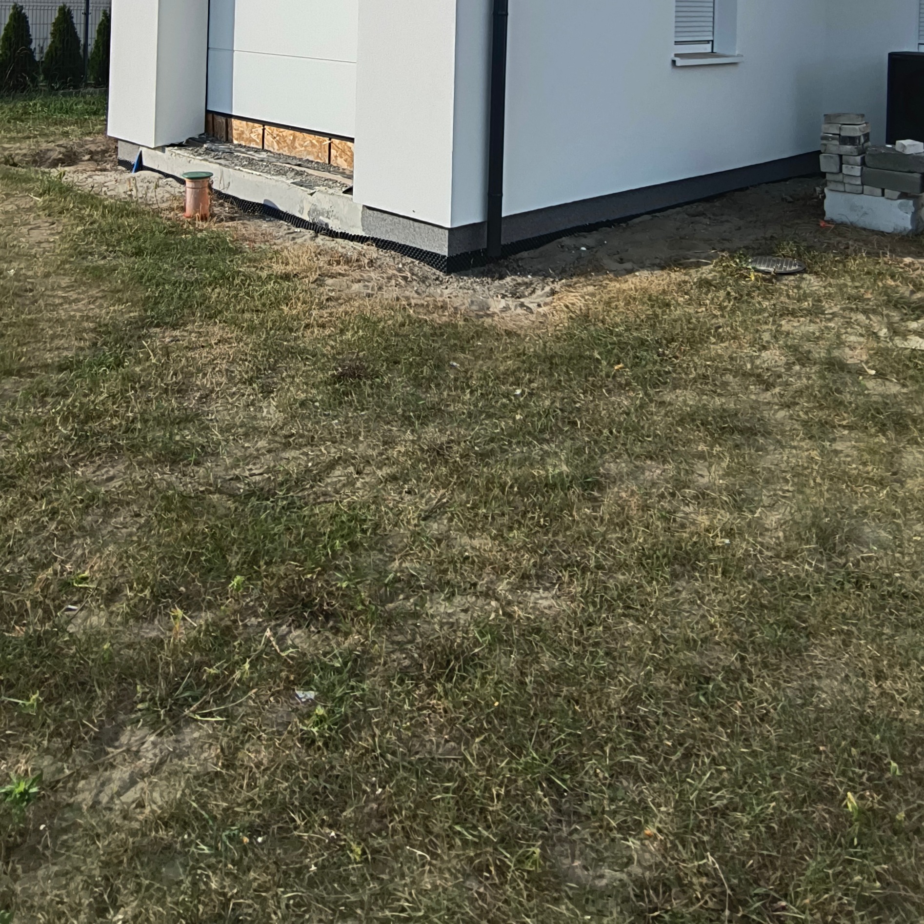 Fragment elewacji budynku z widoczną izolacją fundamentów i czarną rurą drenażową w ziemi. Widoczne cegły i elementy konstrukcyjne.