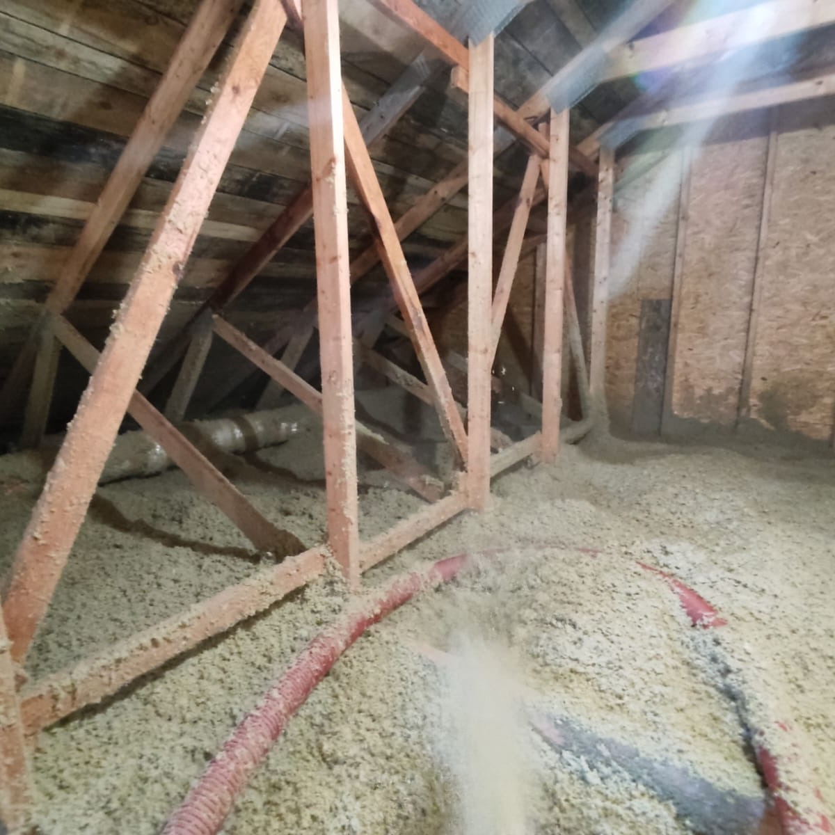 Poddasze z drewnianą konstrukcją wypełniane izolacją wdmuchiwaną z czerwonego węża. Widoczne włókna izolacyjne w powietrzu.