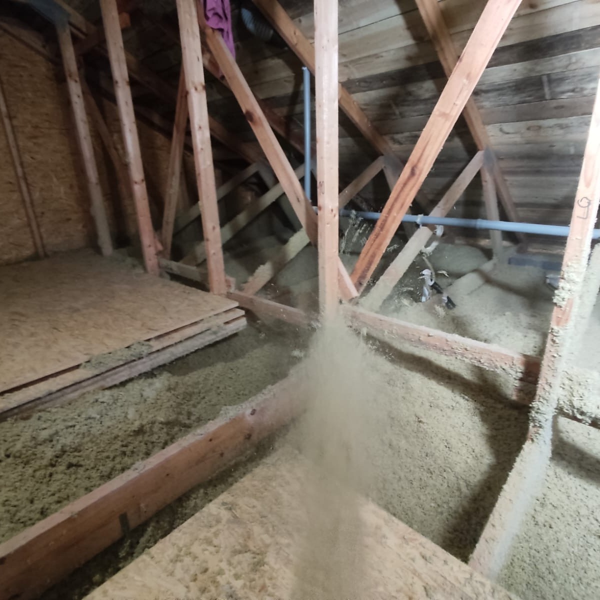 Poddasze z drewnianą konstrukcją, częściowo ocieplone wełną celulozową, z widocznymi płytami OSB. Proces ocieplania w toku, z unoszącym się pyłem.