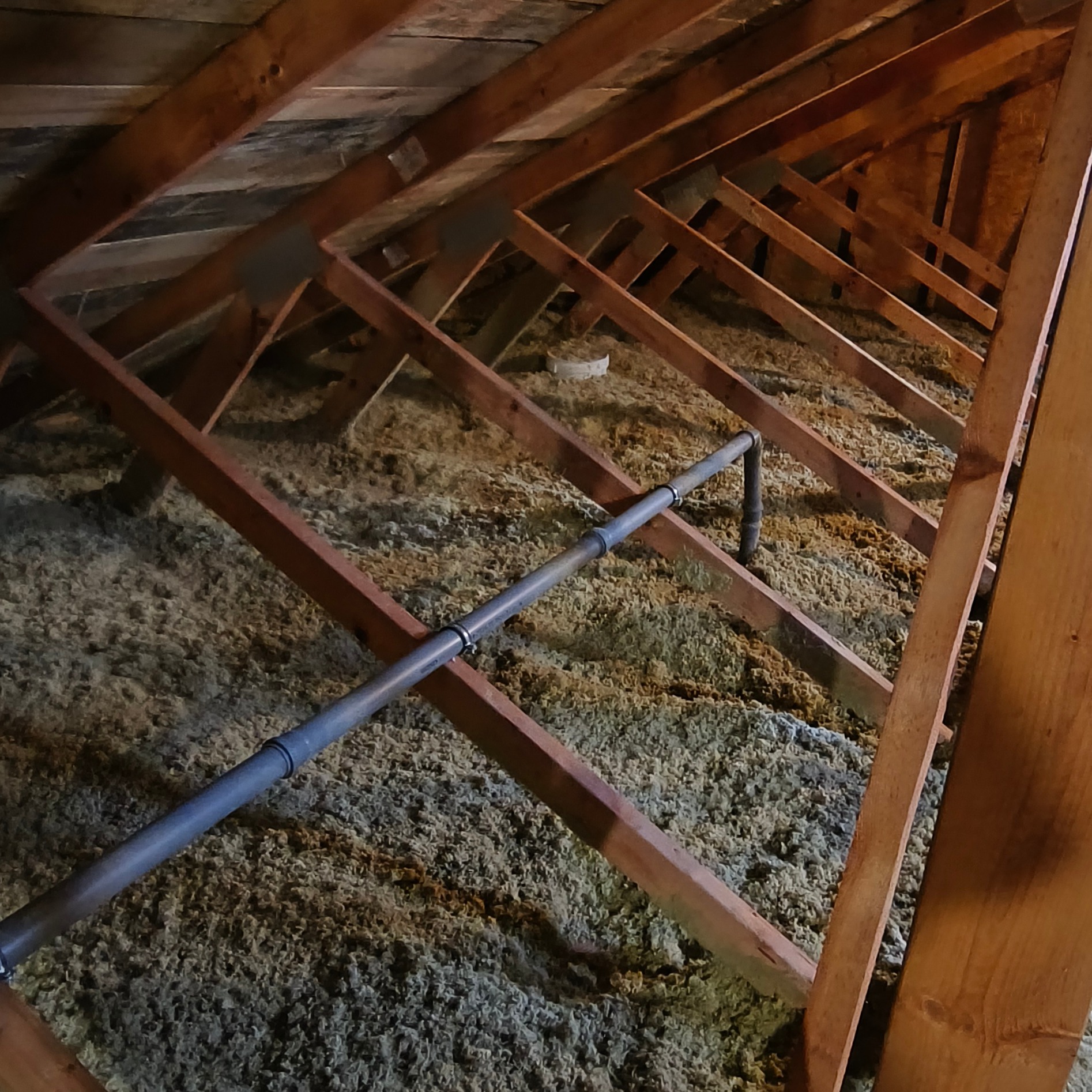 Poddasze z drewnianą konstrukcją i szarą izolacją z wełny mineralnej oraz rurą instalacyjną. Widok z dołu, ukośne ujęcie.