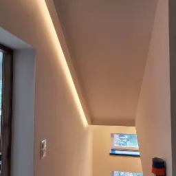 Klatka schodowa po odświeżeniu ścian, z zamontowanym oświetleniem LED wzdłuż sufitu i zabezpieczonymi schodami taśmą malarską.