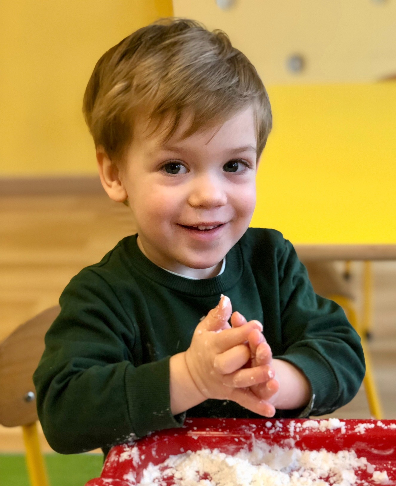 Uśmiechnięty chłopiec z blond włosami w zielonym swetrze, z umązanymi w białej masie rączkami, siedzi przy czerwonym stole na tle żółtej ściany.