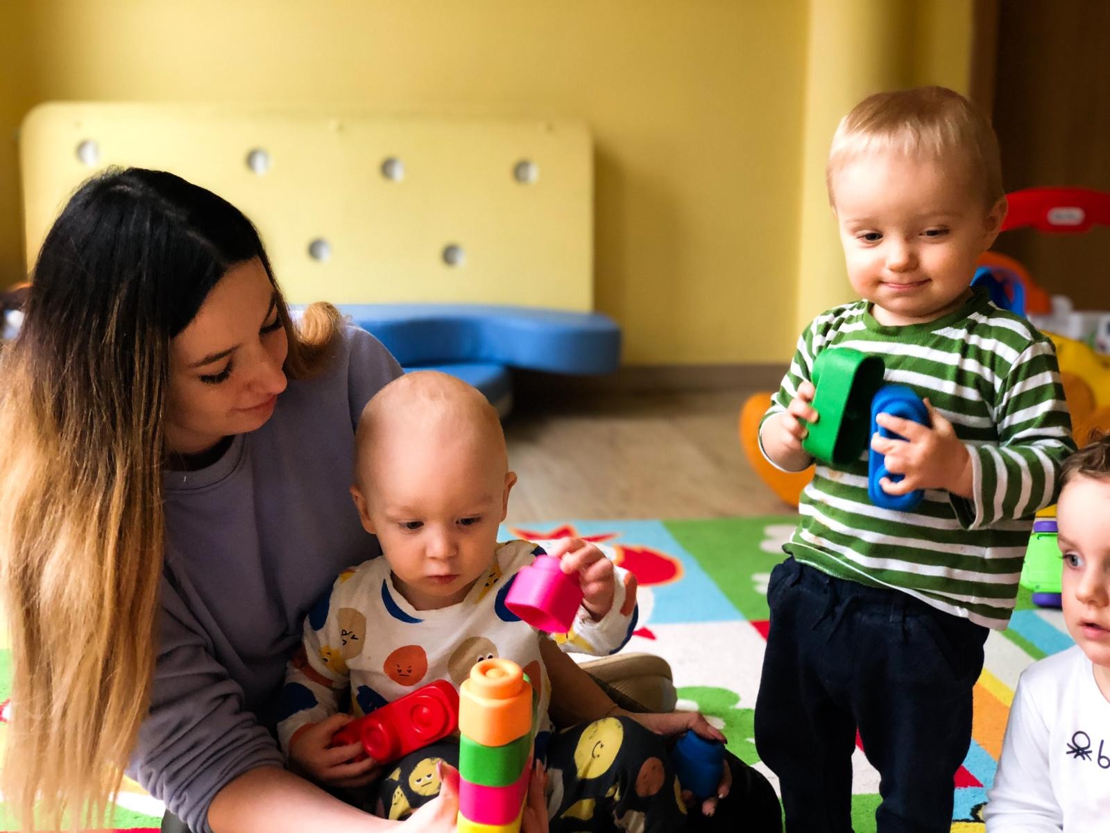 Opiekunka z dziećmi bawi się klockami w sali zabaw w Warszawie. Dziecko w koszulce w emotikony siedzi na kolanach opiekunki i układa wieżę z plastikowych elementów.