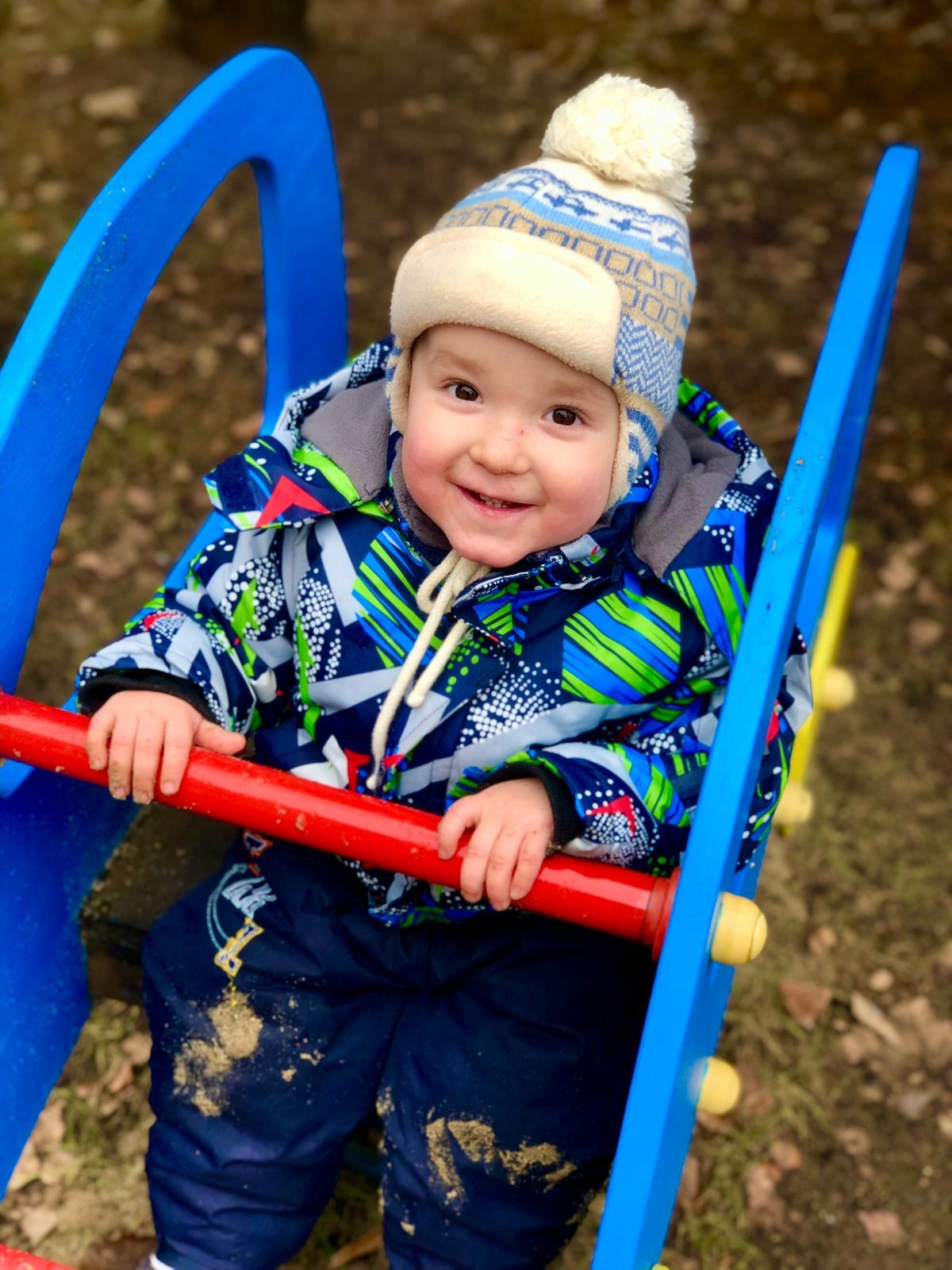 Uśmiechnięty maluch w zimowej czapce i kurtce siedzi na niebiesko-czerwonej konstrukcji, prawdopodobnie na placu zabaw, z piaskiem na spodniach.