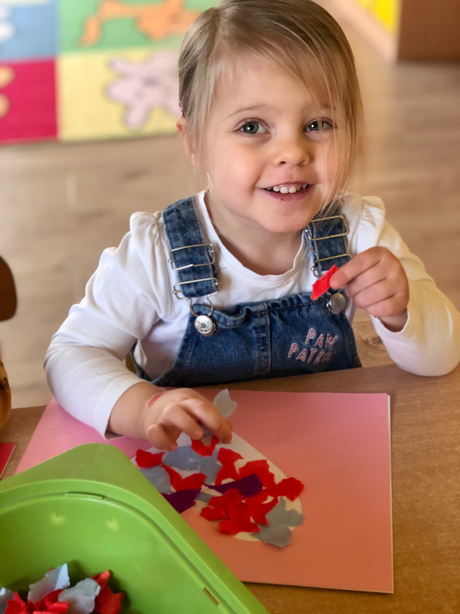 Uśmiechnięta dziewczynka w dżinsowych ogrodniczkach z napisem 'Paw Patrol' tworzy kolaż z kolorowych papierów na różowej kartce, przy zielonym pojemniku z resztkami papieru.
