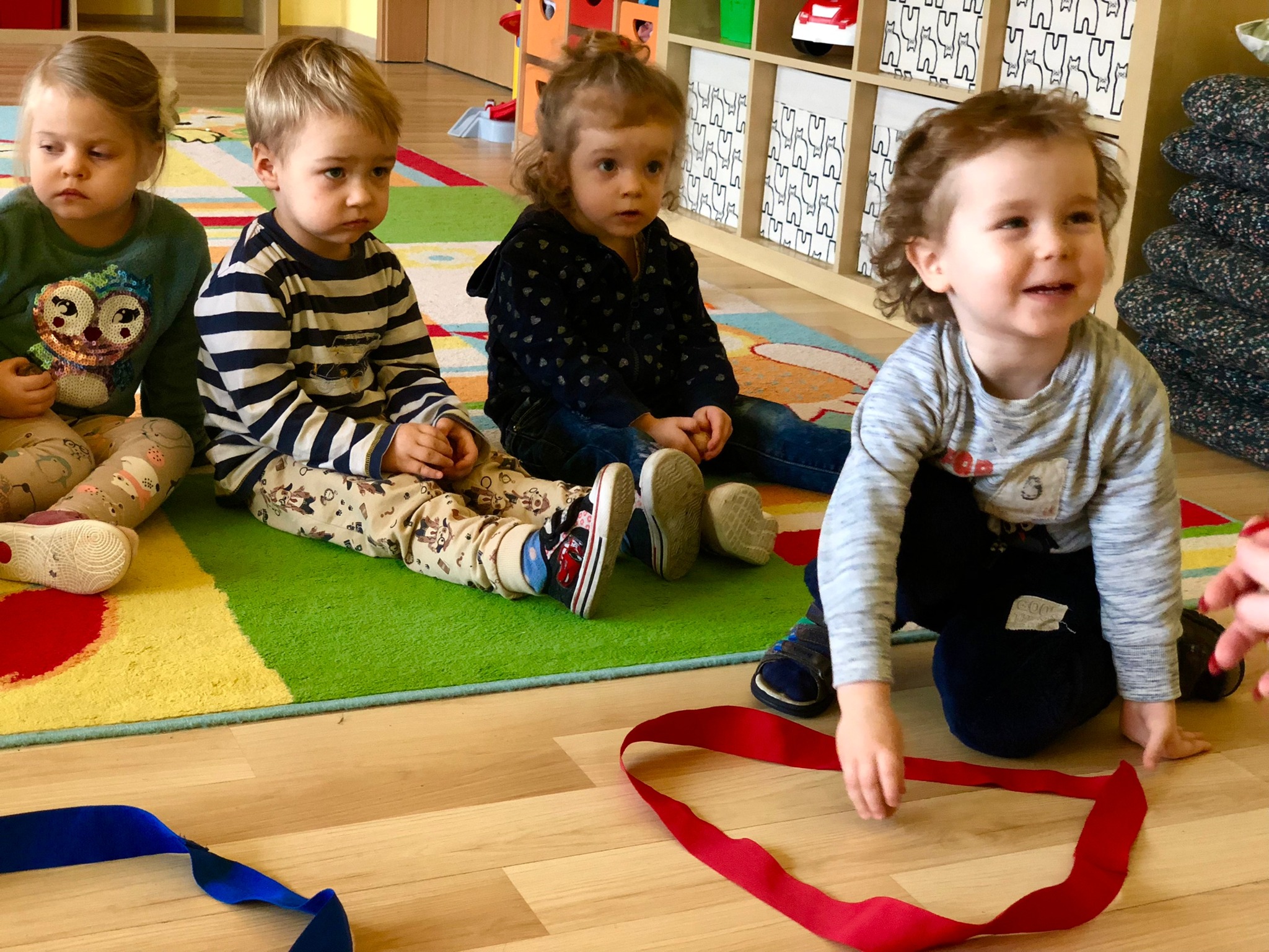 Czwórka dzieci siedzi na podłodze w sali zabaw, jedno z nich bawi się czerwoną wstążką, w tle kolorowy dywan i regał z zabawkami.