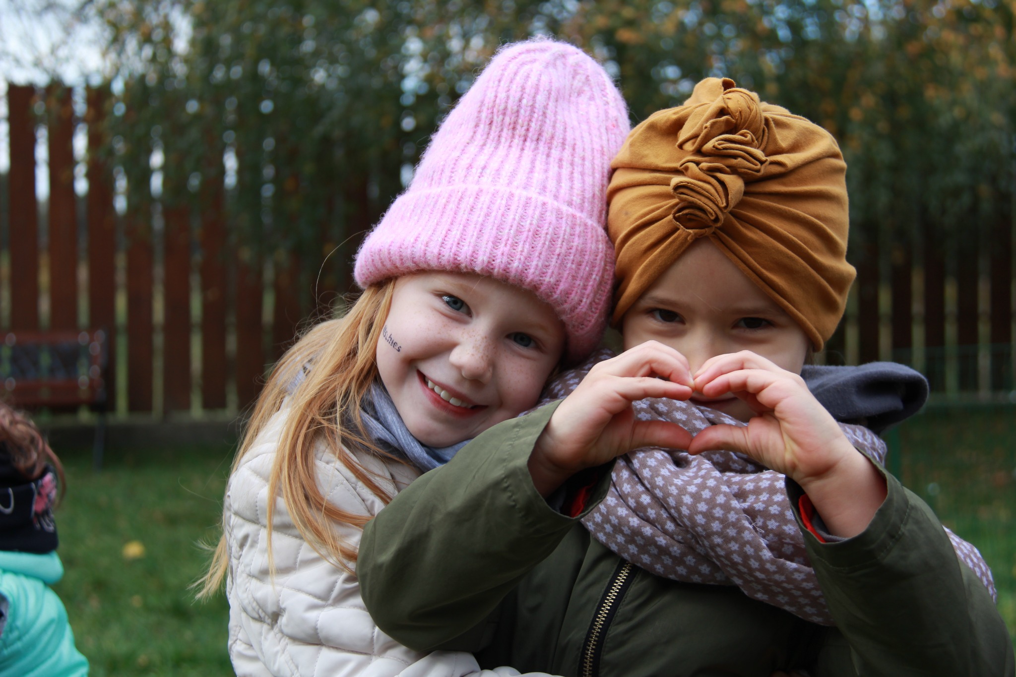 Dwie uśmiechnięte dziewczynki w jesiennych ubraniach, jedna z różową czapką, druga z turbanem na głowie, formują dłońmi serce na tle zieleni.