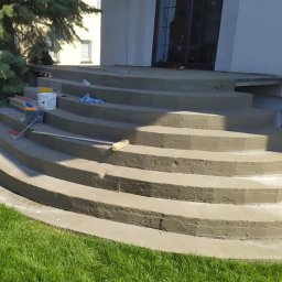 Wykończenia wnętrz - Schody wejściowe do budynku po remoncie, z widocznymi narzędziami malarskimi i wiaderkiem z farbą na jednym ze stopni, otoczone zielenią.