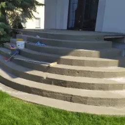 Schody wejściowe do budynku po remoncie, z widocznymi narzędziami malarskimi i wiaderkiem z farbą na jednym ze stopni, otoczone zielenią.