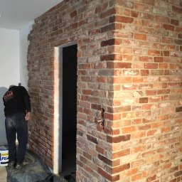 Wykończenia wnętrz - Ściana wykończona cegłą dekoracyjną w odcieniach czerwieni i brązu, widoczny fragment pracownika w ubraniu roboczym i wiadro farby, przejście do innego pomieszczenia w tle.