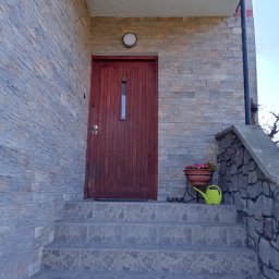 Wykończenia wnętrz - Wejście do domu z drewnianymi drzwiami z numerem 25, wykończone kamieniem elewacyjnym w odcieniach szarości, schody wyłożone płytkami, donica z kwiatami i zielona konewka.