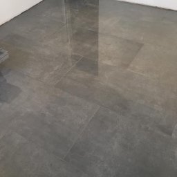 Wykończenia wnętrz - Ułożone, szare, matowe płytki podłogowe z widocznym odbiciem światła, świeżo po umyciu w Stargardzie, z mopem i wiadrem w tle.
