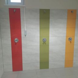 Wykończenia wnętrz - Trzy kabiny prysznicowe wykończone jasnymi płytkami z pionowymi pasami w kolorach: czerwonym, zielonym i pomarańczowym. Każda kabina wyposażona w chromowaną baterię i wieszak.