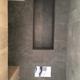 Wykończenia wnętrz - Szara ściana łazienki z marmurowymi płytkami, wnęką na kosmetyki wyłożoną tym samym materiałem oraz chromowanym przyciskiem spłuczki, z odbiciem osoby robiącej zdjęcie.