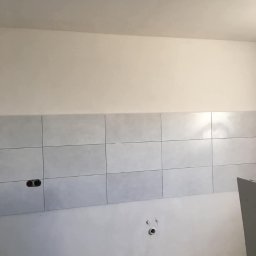 Wykończenia wnętrz - Ściana w trakcie układania jasnoszarych płytek, widoczne puszki elektryczne i instalacja wodna, surowy stan wykończenia.